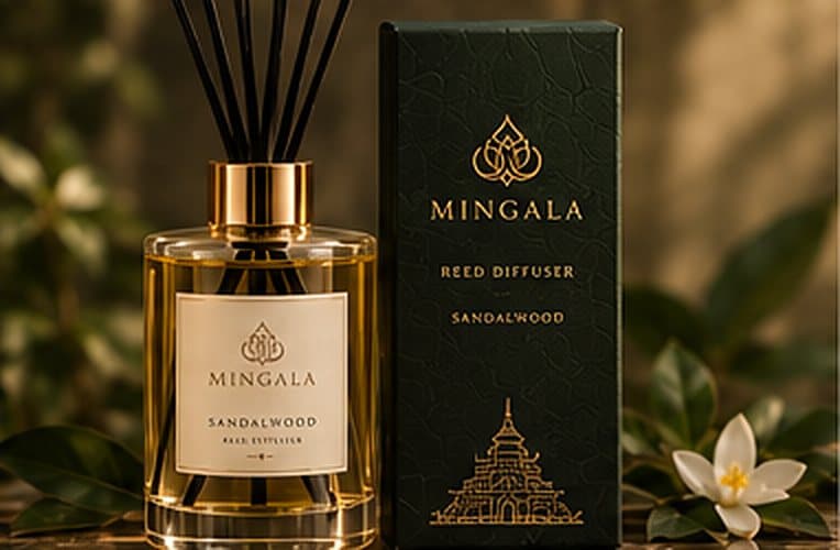 A glass reed diffuser with black reeds in front of a dark green MINGALA box